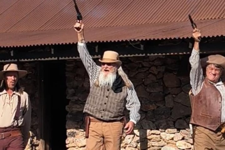 Three men in cowboy outfits raising pistols outdoors against a rustic backdrop.