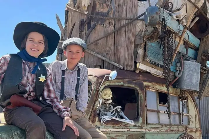 Two children dressed in old-fashioned attire sit on a vintage car with a skeleton and piled items.