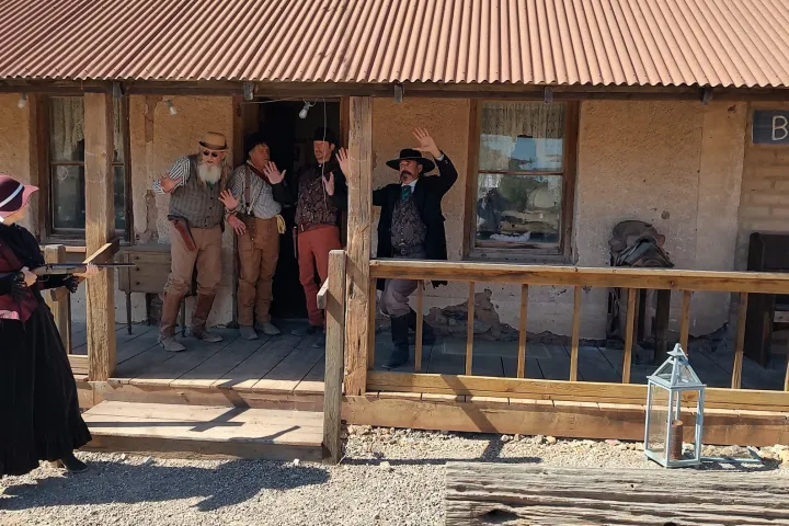 Woman aims rifle at four men with hands raised on wooden porch of rustic building.