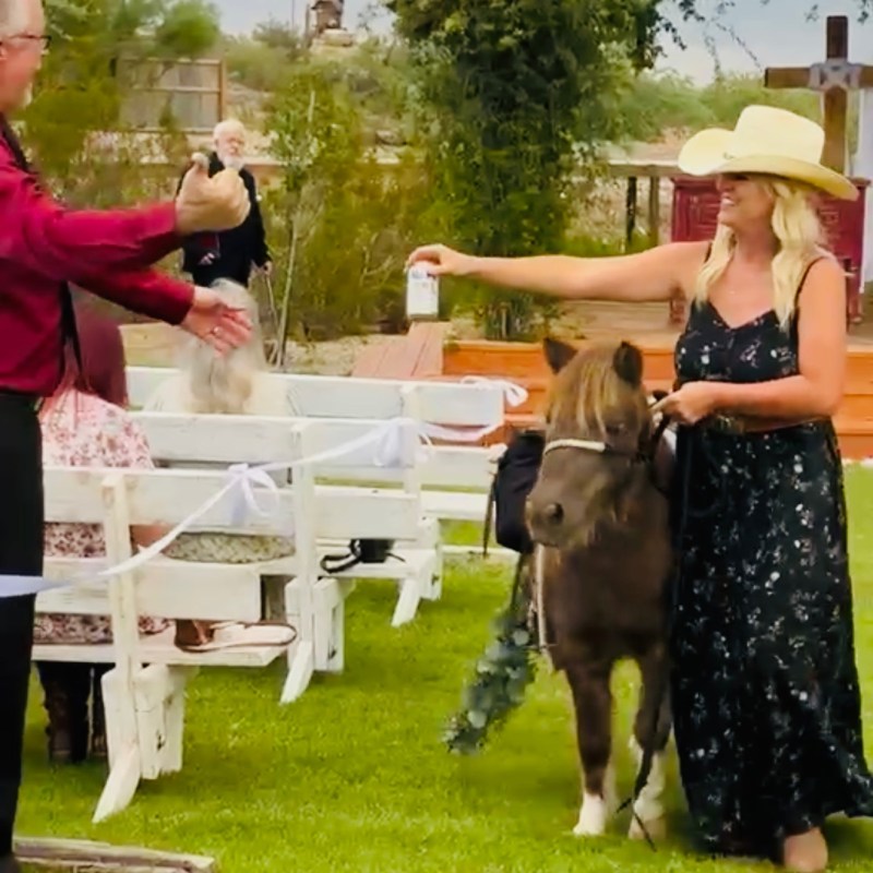 Woman in hat with pony hands drink to man at outdoor event.