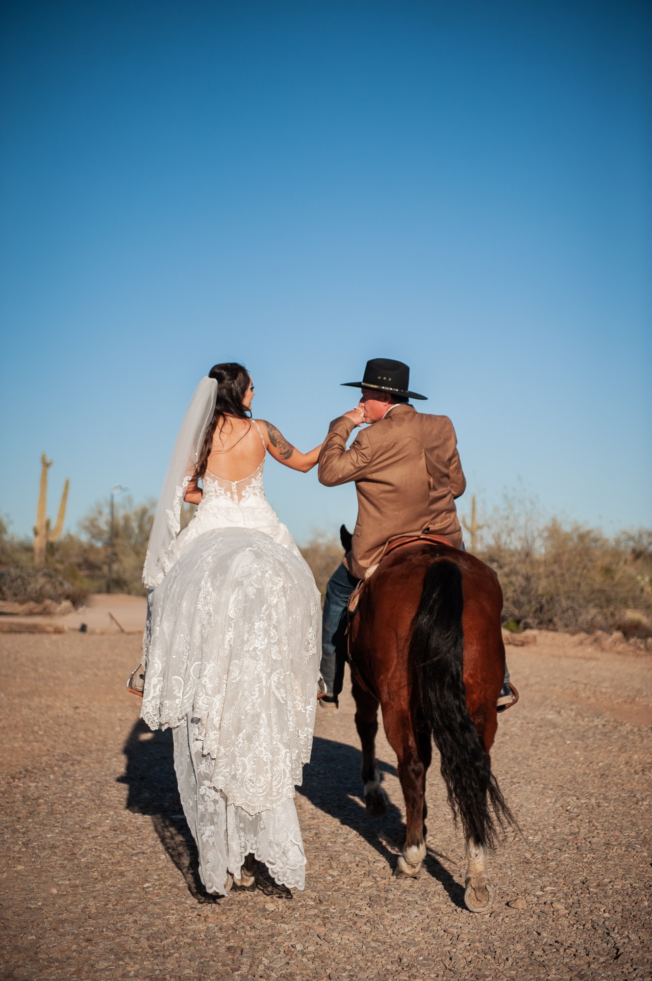 Bride and cowboy groom on horseback in desert, holding hands under clear blue sky.